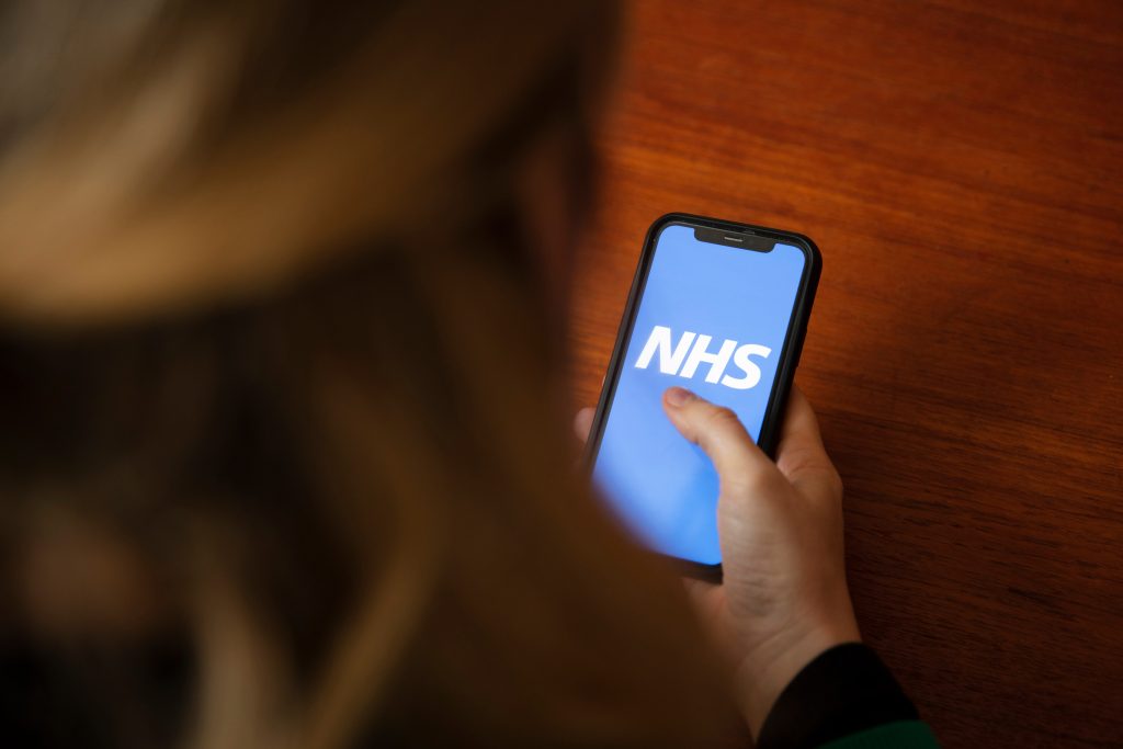 Close-up of a person holding a smartphone displaying the bright blue NHS app logo on the screen. The phone is held over a polished wooden table, with the person’s blond hair slightly out of focus in the foreground.