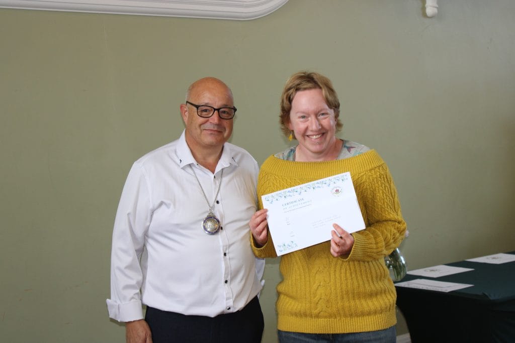 A man in a white shirt and glasses stands beside a smiling woman in a yellow jumper, who is holding a certificate of achievement. They are indoors, against a plain light green wall.