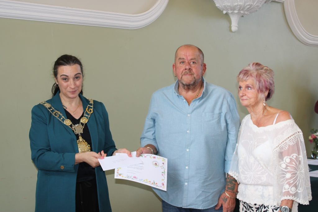 A woman in ceremonial chains and a teal jacket hands a certificate to a man in a pale blue shirt, who stands beside an older woman with light purple hair wearing a white lace top. All three are indoors, with a light green wall in the background.
