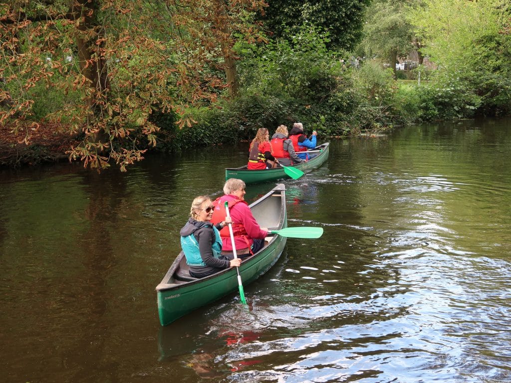 People paddling in green canoes along a calm river, surrounded by leafy trees and greenery. The group wears life jackets and outdoor clothing, with one woman looking back and smiling at the camera.