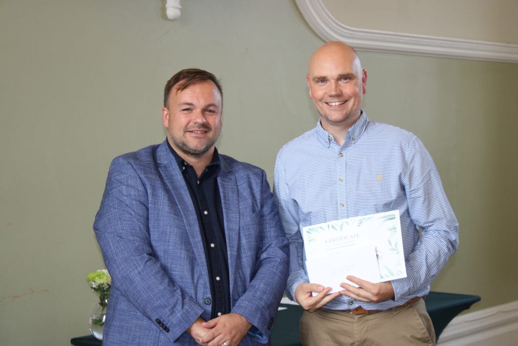 Two men standing side by side indoors. One wears a pale blue striped shirt and holds a certificate of achievement; the other wears a navy shirt and blue blazer. Both are smiling at the camera.