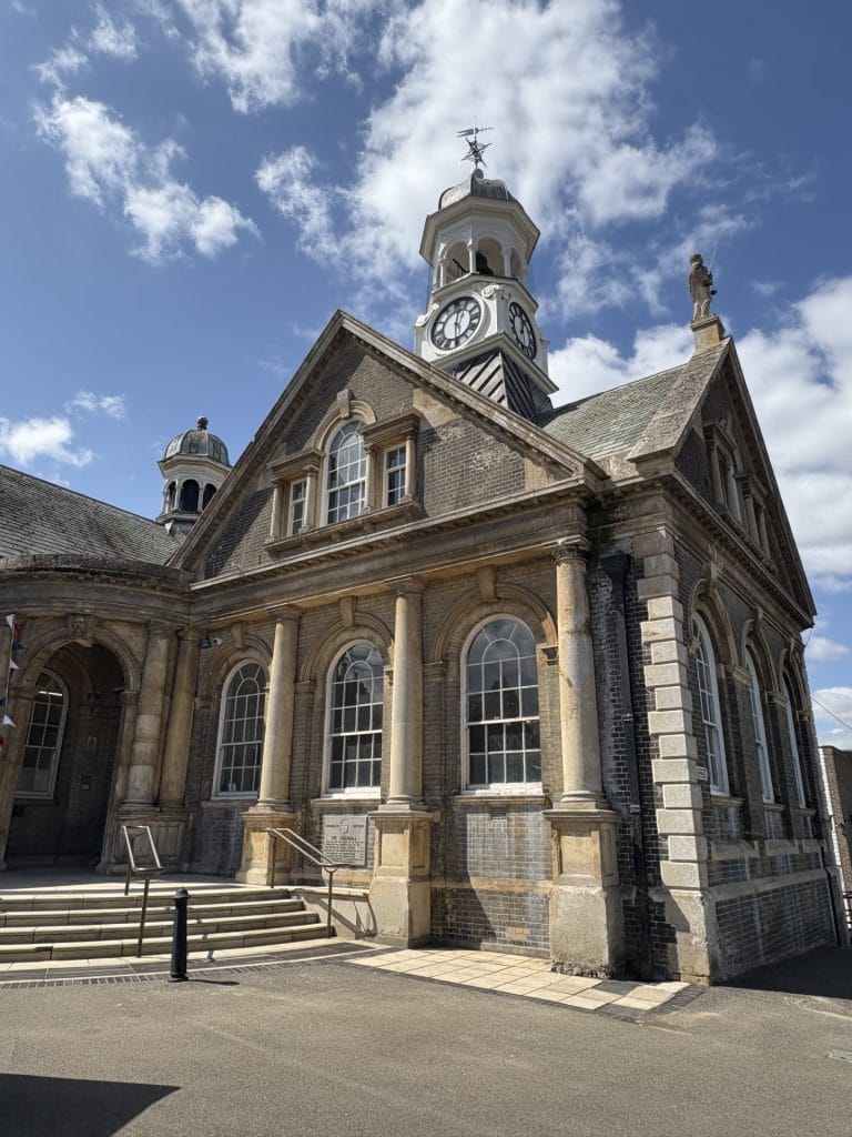 View of the front of Thetford Guildhall under a bright blue sky with scattered clouds. The historic building features arched windows, stone columns, and a central clock tower with a white-painted cupola and weather vane. A restored black and white clock face is clearly visible on the tower.