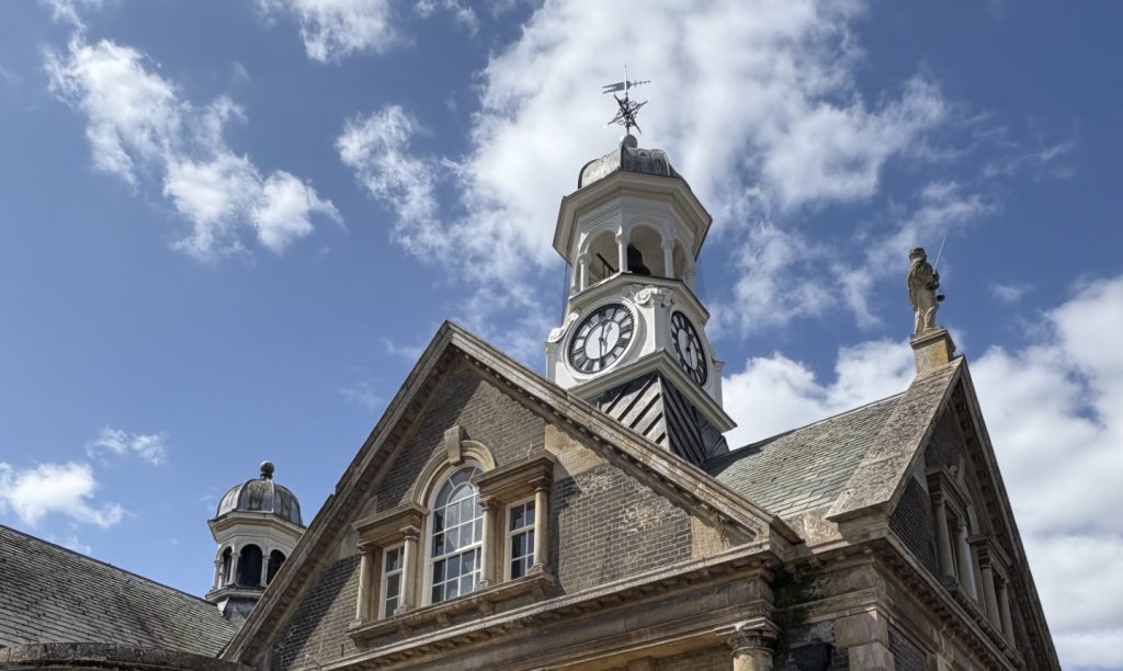 View of the front of Thetford Guildhall under a bright blue sky with scattered clouds. The historic building features arched windows, stone columns, and a central clock tower with a white-painted cupola and weather vane. A restored black and white clock face is clearly visible on the tower.