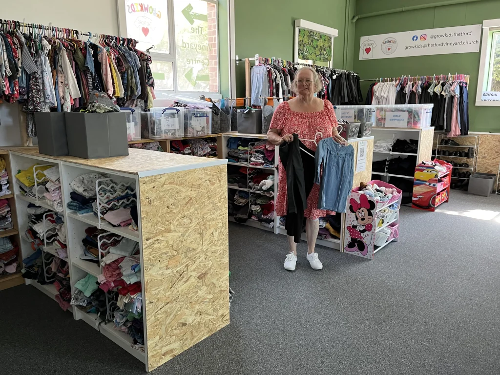 A smiling woman in a red dress holds up two children’s outfits in a bright and neatly organised room full of donated clothes. The space is part of Thetford Vineyard Church’s Growkids initiative, offering free clothing for children. Behind her are racks of children’s clothing sorted by size, plastic tubs on shelves, and a corner with children’s shoes and toys. A large window and green painted walls make the space feel welcoming. On the wall is a banner with logos and social media handles: “@growkidsthetford” and “growkids@thetfordvineyard.church”.
