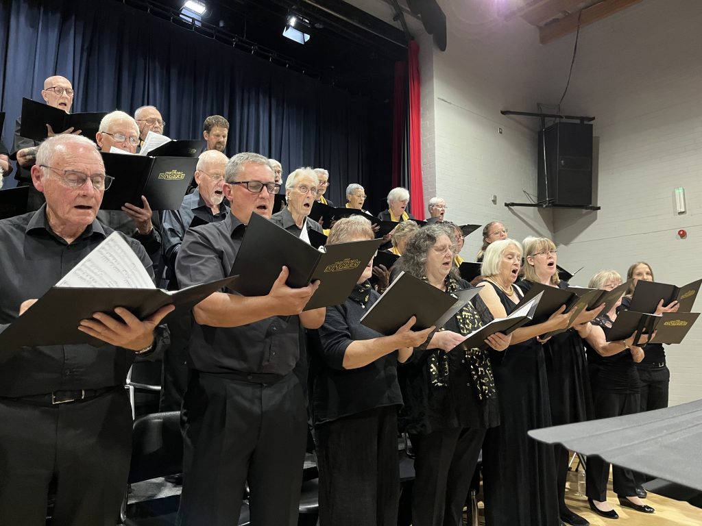 Members of the Thetford Singers choir, dressed in black, sing together from sheet music on a tiered stage, with men and women filling the rows under stage lighting.