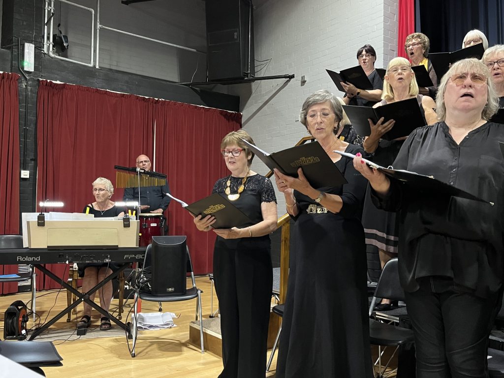 A pianist accompanies the Thetford Singers as women in black outfits perform with sheet music, standing in front of red curtains. A percussionist plays chimes in the background.