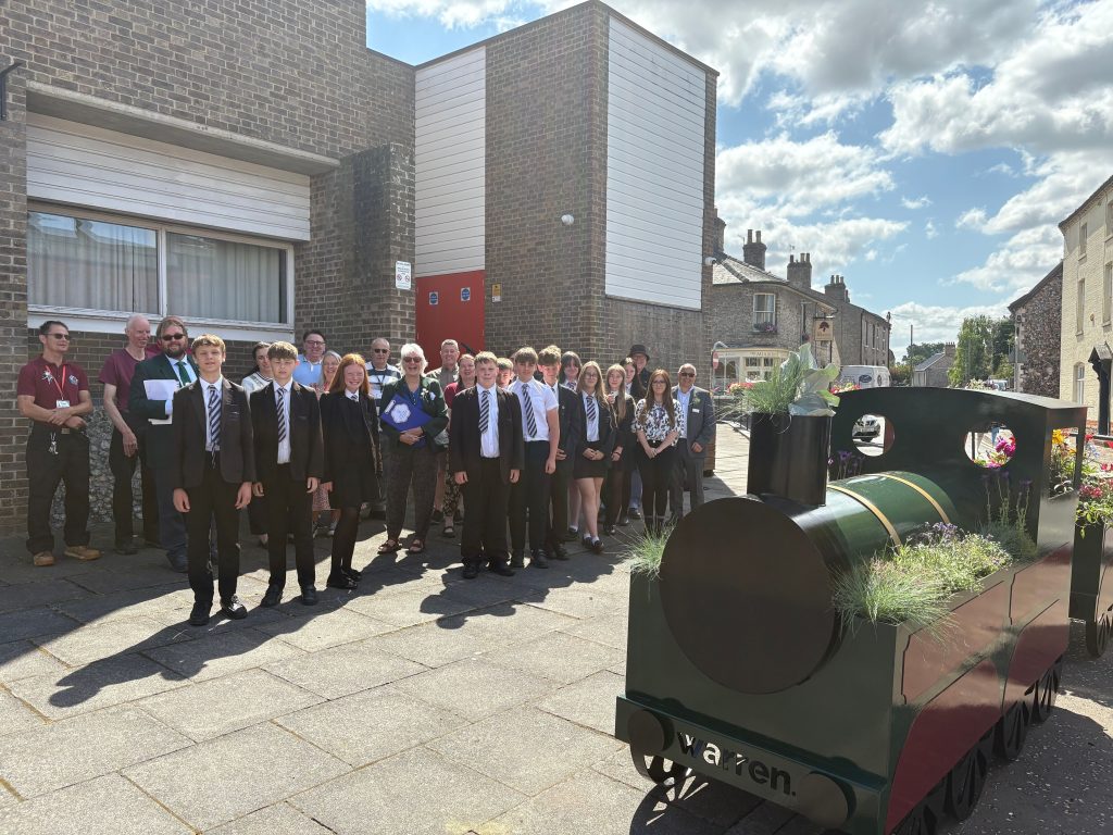 Large group of Thetford Academy students in uniform standing with local representatives and Anglia in Bloom judges beside a green train-shaped planter filled with flowers. The scene is set in Thetford town centre with brick buildings and a blue sky in the background.