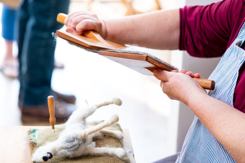 Close-up of a person in a striped apron using two wooden carding paddles to work wool. On the table below is a partially made felted animal figure, with a felting needle resting beside it.