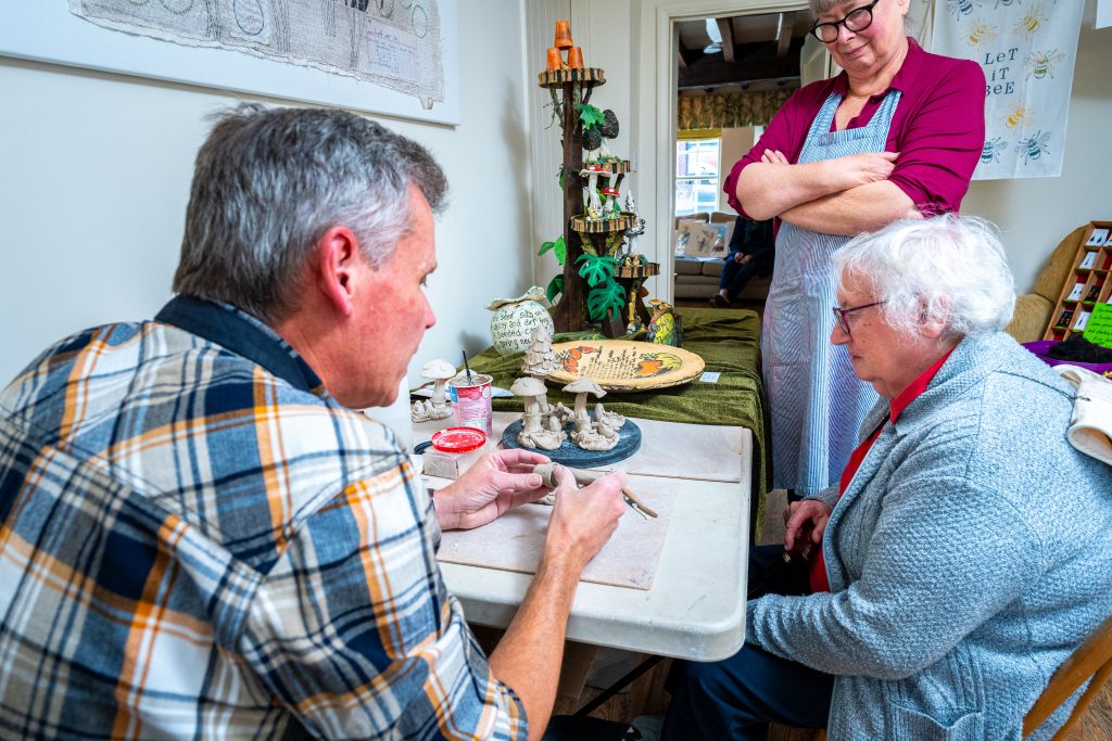 A man in a checked shirt sits at a table sculpting a small clay model of mushrooms while an older woman in a red top and grey cardigan watches closely. A second person in an apron stands with arms folded, observing. Handcrafted pottery and decorative pieces are displayed on a table behind them.