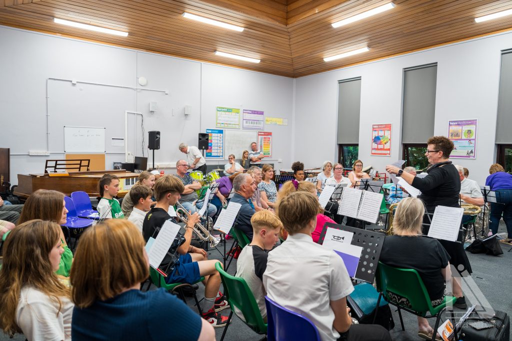 Members of Breckland Brass Band rehearse in a bright hall, with children and adults playing brass instruments under the direction of a conductor.