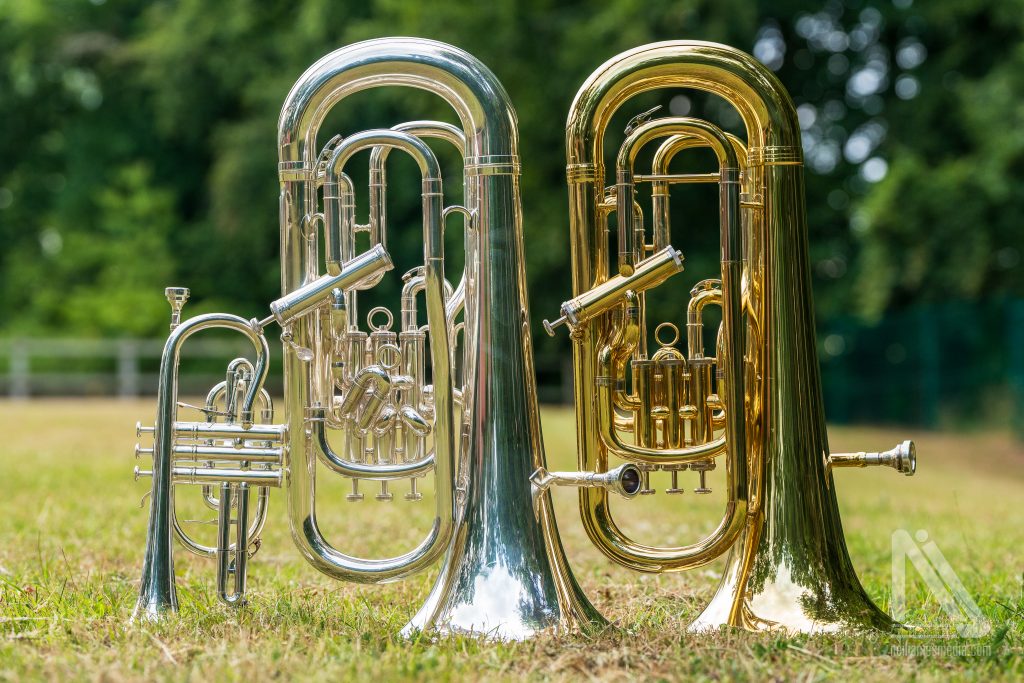 Three polished brass instruments from Breckland Brass Band, two gold and one silver, stand on the grass outdoors, gleaming in the sunlight.