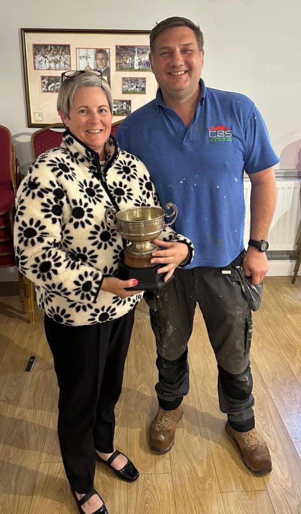 A smiling woman in a black-and-white floral fleece holds a large silver trophy beside a man in a blue work polo shirt with paint marks.
