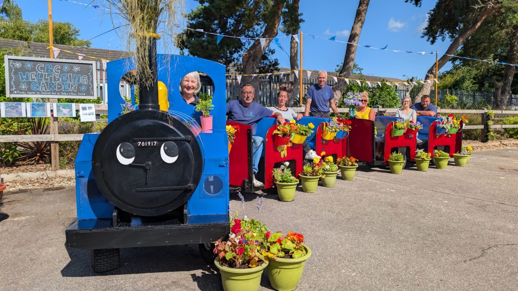 Group of people smiling while sitting in a brightly painted blue and red wooden train planter decorated with colourful flowers in pots. Behind them is a sign reading "CBC Well-being Garden" with bunting and trees in the background.