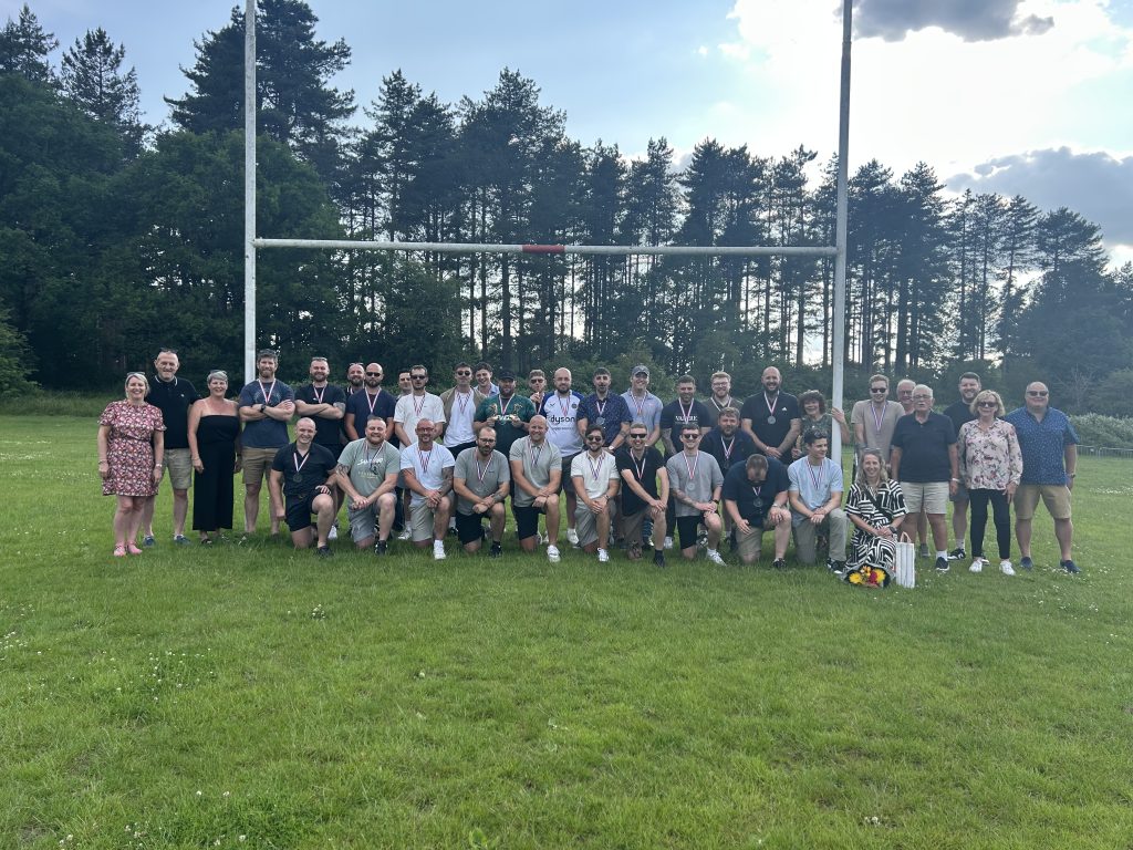 A large group photo of Thetford rugby players, coaches, and supporters standing under rugby goalposts on a sunny field, many wearing medals.