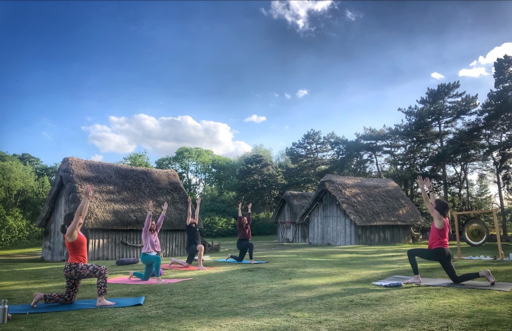 A group of five people take part in an outdoor yoga session on a grassy lawn beside reconstructed Anglo-Saxon buildings at West Stow Anglo-Saxon Village. Participants are practising a lunge pose with their arms raised towards the sky on colourful yoga mats. The evening scene is framed by thatched buildings, pine trees and a bright blue sky with scattered clouds, creating a peaceful countryside setting.