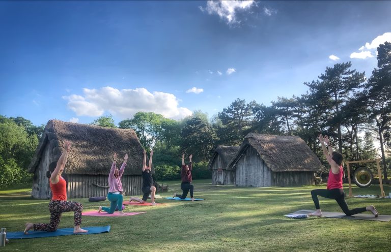A group of five people take part in an outdoor yoga session on a grassy lawn beside reconstructed Anglo-Saxon buildings at West Stow Anglo-Saxon Village. Participants are practising a lunge pose with their arms raised towards the sky on colourful yoga mats. The evening scene is framed by thatched buildings, pine trees and a bright blue sky with scattered clouds, creating a peaceful countryside setting.