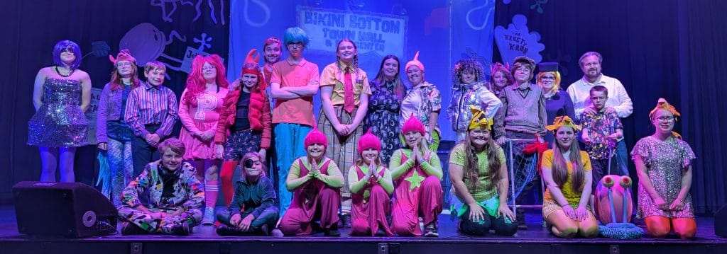 A colourful group photo of children and adults dressed in creative, themed costumes on a stage set inspired by SpongeBob SquarePants. The cast is smiling and gathered in rows, with props and stage decoration behind them, including signs reading “Bikini Bottom Town Hall” and “The Krusty Krab”.