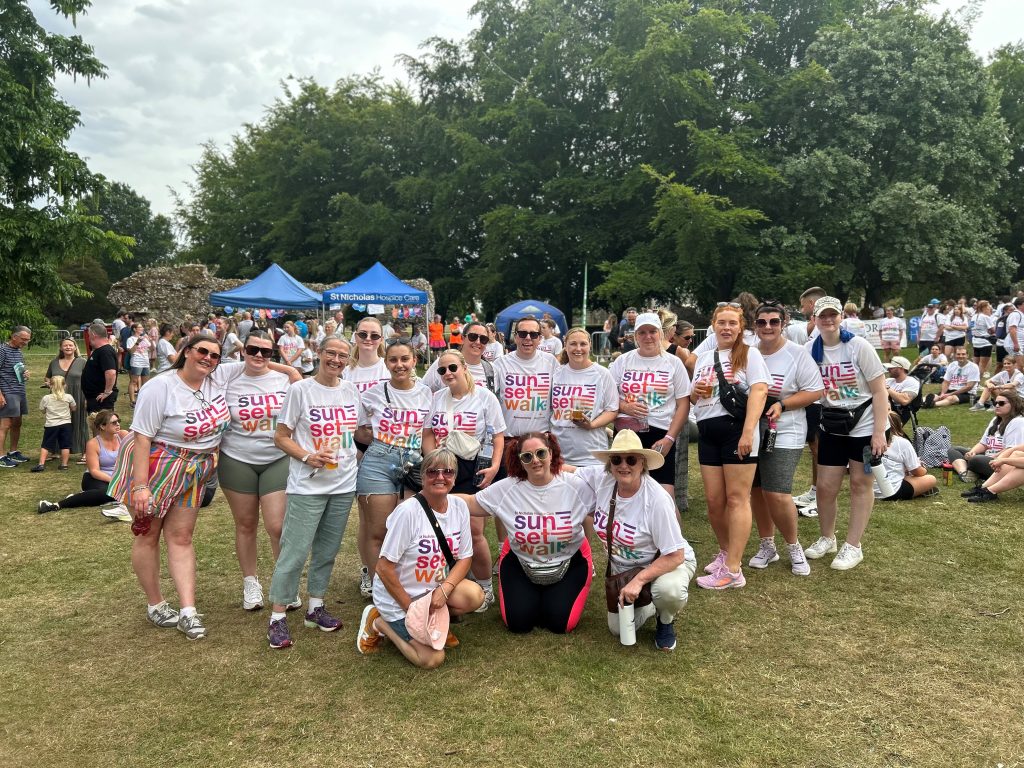 A large group of smiling walkers wearing matching white "Sunset Walk" T-shirts gather together on grass in front of blue St Nicholas Hospice Care tents. More participants and spectators can be seen sitting and standing in the background under tall green trees.