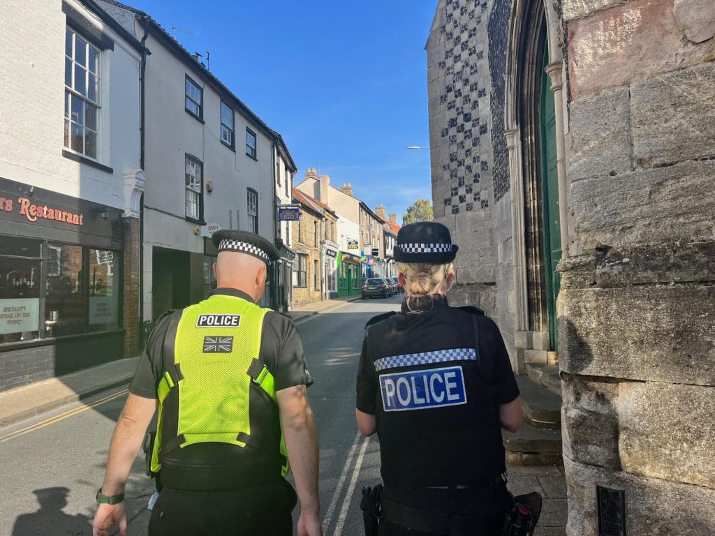 Two police officers, one male and one female, walk down Whitehart Street in Thetford on a sunny day. They are seen from behind, with the male officer wearing a bright yellow vest and the female officer in a black uniform. To the left is the exterior of Cheers Restaurant, and to the right is the stone entrance of a historic building with flint and stonework. The street ahead is lined with shops and terraced buildings.