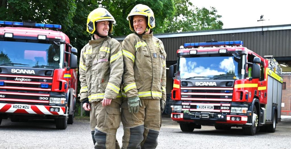 Two firefighters in full uniform and helmets stand back-to-back, smiling at each other outside a fire station. Behind them are two red Scania fire engines with blue lights, parked on either side of the driveway. The atmosphere is bright and friendly, with greenery and station buildings in the background.