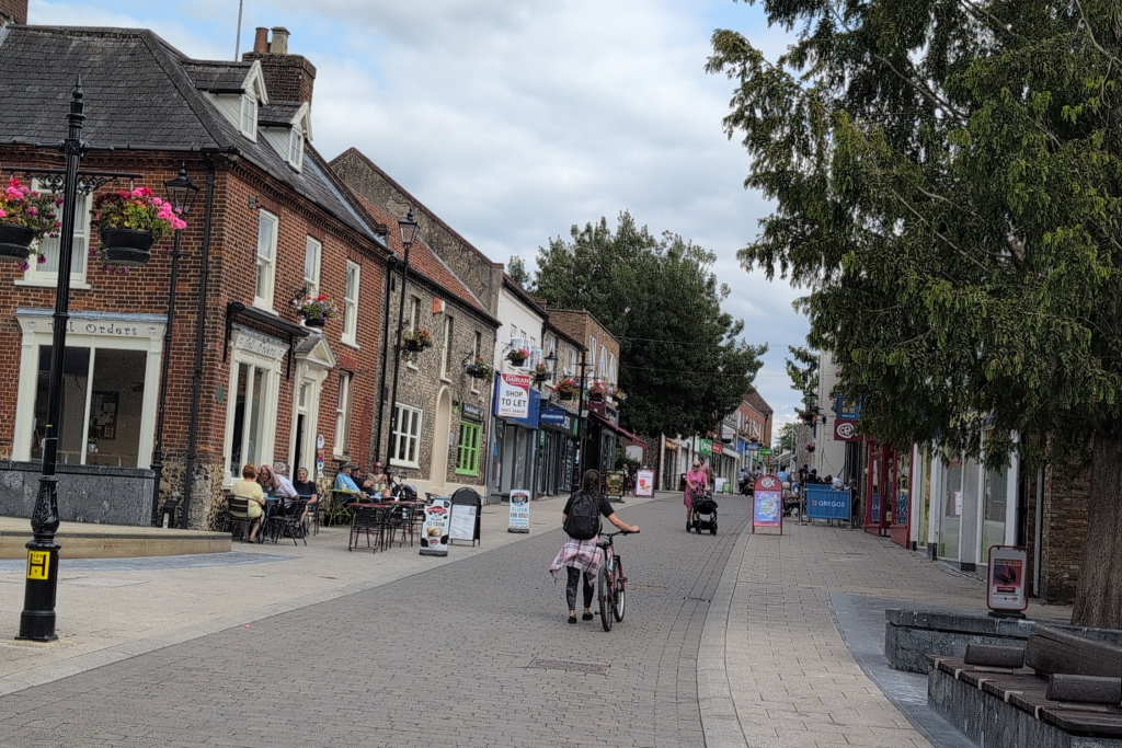 A view of Thetford town centre showing shops and cafés on a cobbled street with hanging flower baskets. A person pushes a bicycle along the street while others sit at tables outside and people walk further up the street.