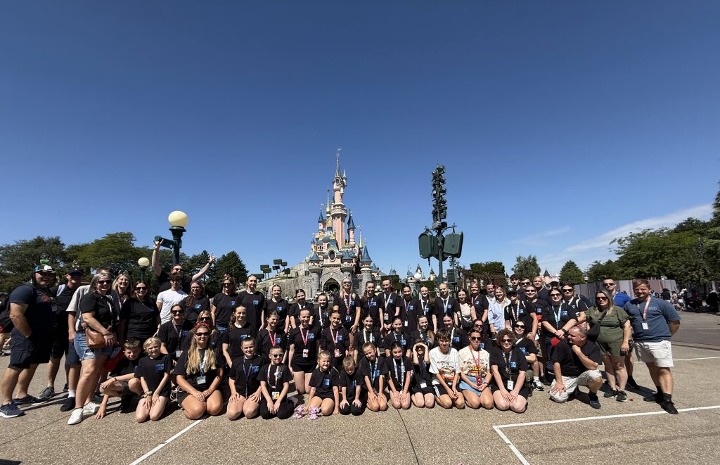 A large group of dancers, children, and adults pose together in front of the fairytale-style castle at Disneyland Paris. The group is smiling and wearing matching black t-shirts under a bright blue sky.