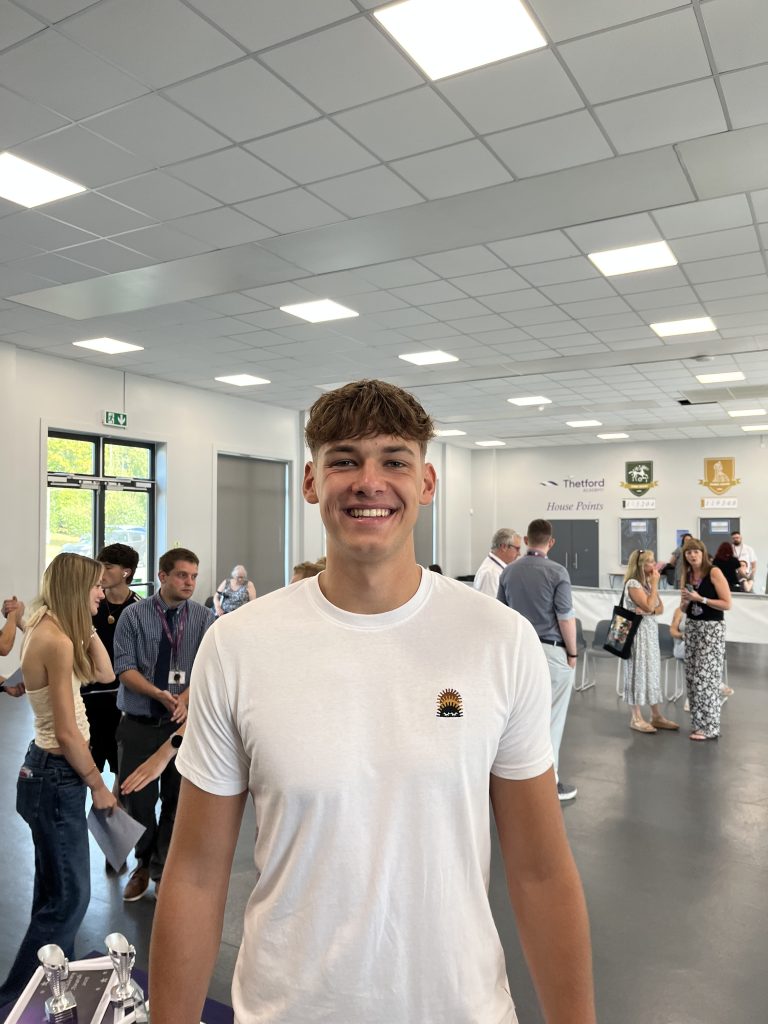 A smiling student in a white t-shirt stands in the foreground at Thetford Academy, with classmates and teachers visible in the background.