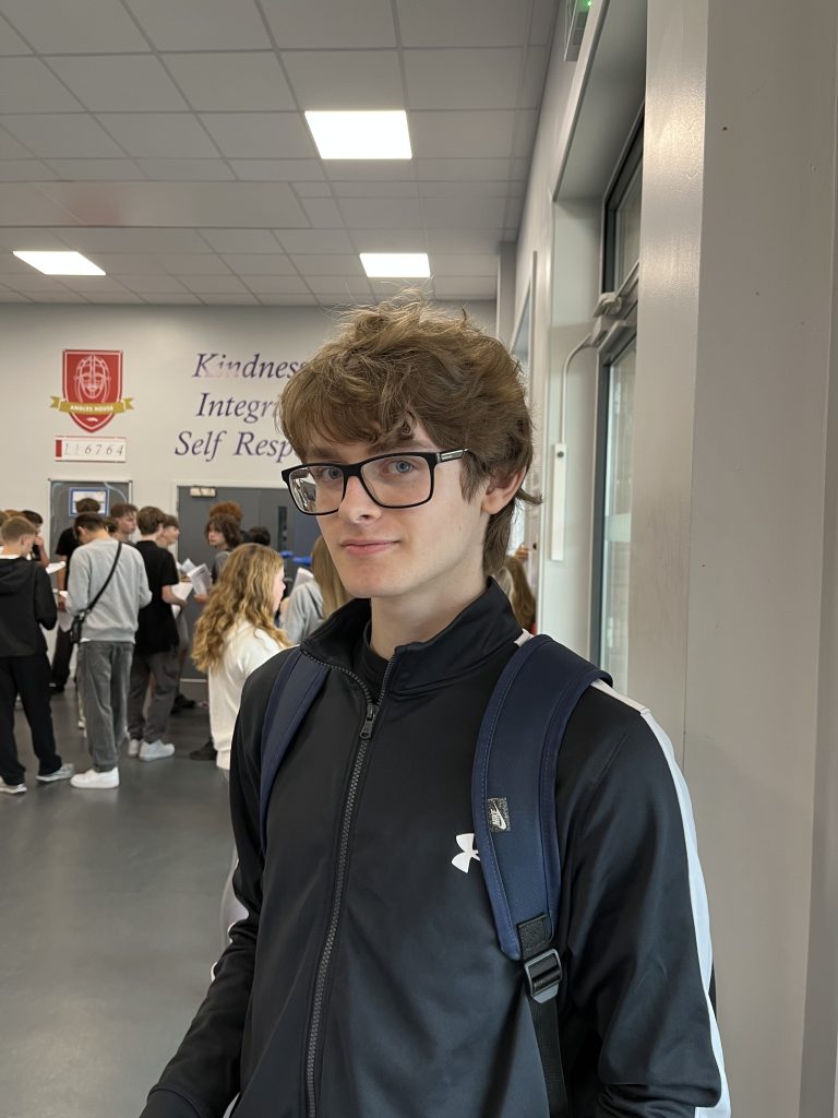 A student wearing glasses, a dark zip-up jacket, and a backpack stands indoors at Thetford Academy, with other students gathered in the background.