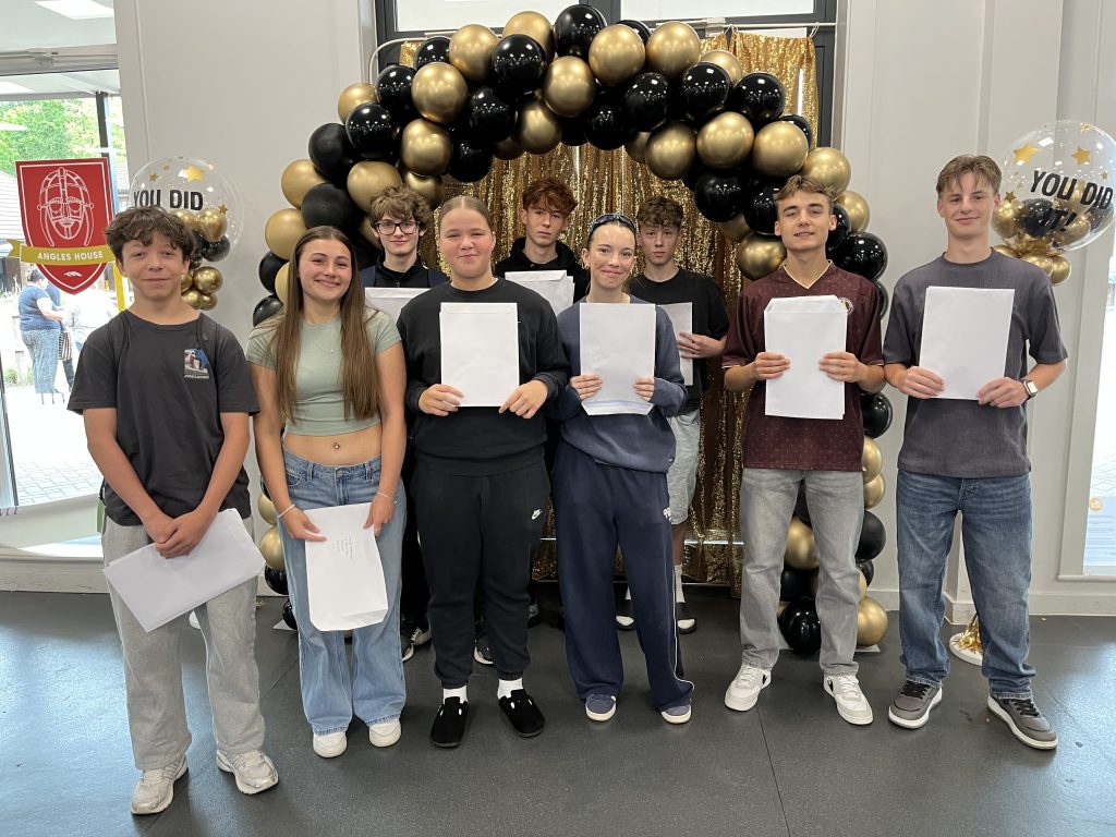 A group of eight smiling students stand in front of a black and gold balloon arch, each holding their GCSE results envelopes. Decorative balloons read “You Did It!”.
