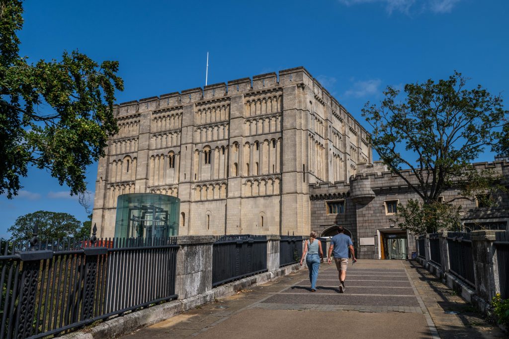 Two people walking towards Norwich Castle, a large Norman stone fortress with rows of arched windows and battlements, set against a clear blue sky. A modern glass lift structure is visible on the left, and trees and railings frame the walkway leading to the entrance.