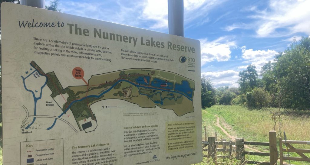 Entrance sign at The Nunnery Lakes Reserve with a map and visitor information, showing footpaths, observation hides, and wildlife habitats. The board is titled “Welcome to The Nunnery Lakes Reserve” and includes a “You are here” marker. Behind the sign is a grassy footpath with a wooden gate, leading into the reserve under a partly cloudy sky.
