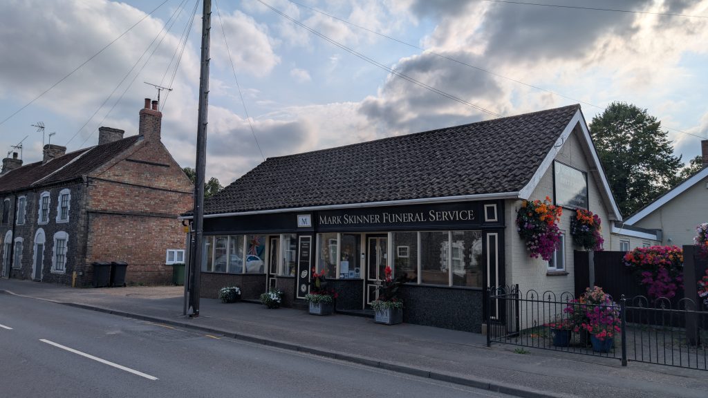 Street view of Mark Skinner Funeral Service, a single-storey building with a tiled roof and black sign above the windows. The frontage is decorated with planters of red, white, and pink flowers, with additional hanging baskets. To the left are older brick terraced houses, and to the right is a light-colored building. A tall telegraph pole and overhead wires cross the sky above.