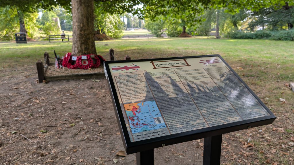 Information board titled “Thetford’s FEPOW Tree” with text and maps explaining the Far East Campaigns during World War II. In the background, a tree with red poppy wreaths at its base is visible, enclosed by chains and posts. The setting is a grassy park with benches and trees.