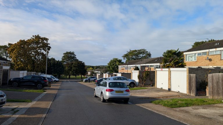 Suburban street lined with parked cars and rows of garages, with terraced houses and trees in the background under a partly cloudy sky.