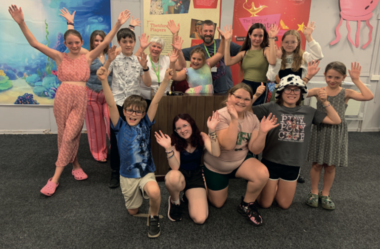 Group of smiling children and a few adults posing indoors with their arms raised in celebration. They are standing and kneeling in front of colourful backdrops and banners, including ones for “Thetford Players” and “The Story of Panto.”