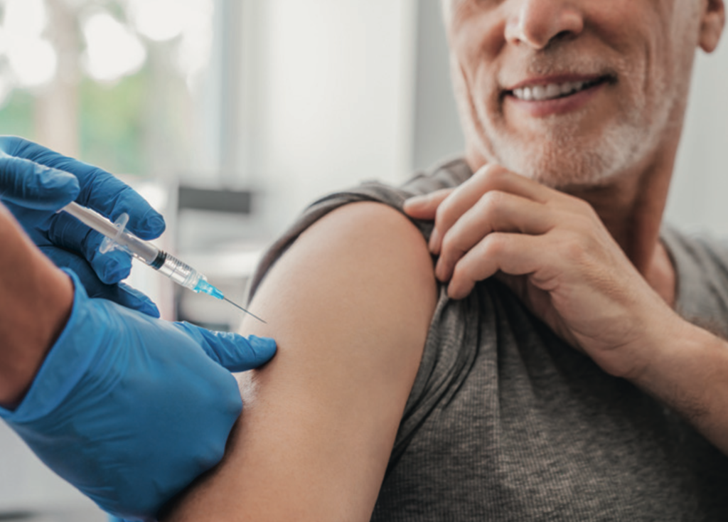 Close-up of a person receiving an injection in their upper arm. A healthcare professional wearing blue gloves holds a syringe, while the patient, smiling, pulls up the sleeve of a grey shirt to expose their arm.