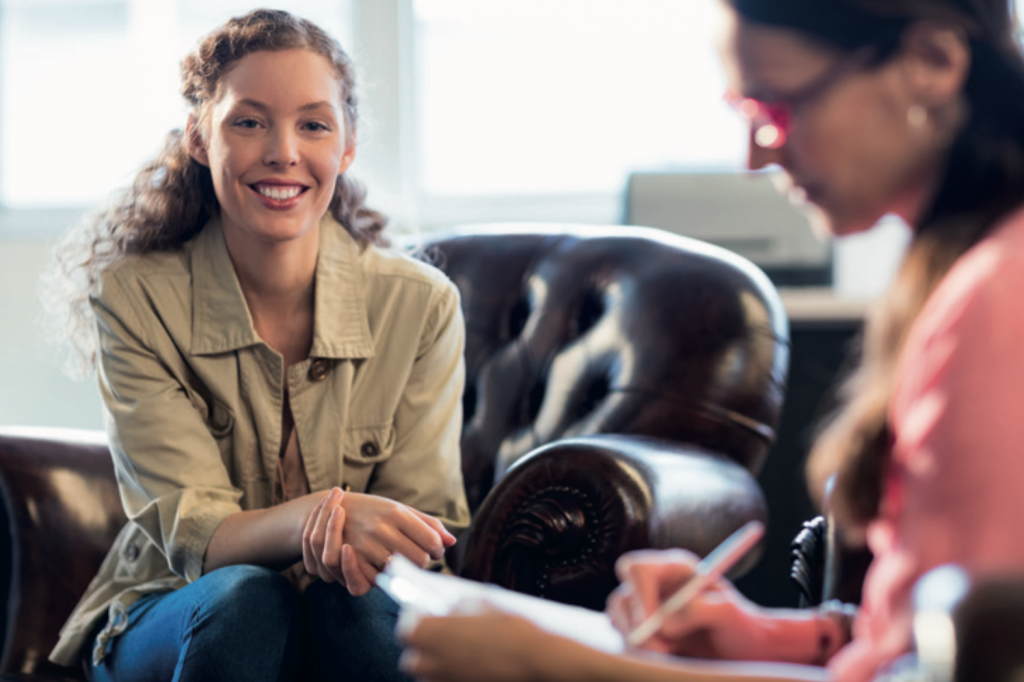 A young woman smiling while sitting on a leather armchair, facing another person who is blurred in the foreground and writing on a clipboard. The setting suggests a relaxed conversation or counselling session.