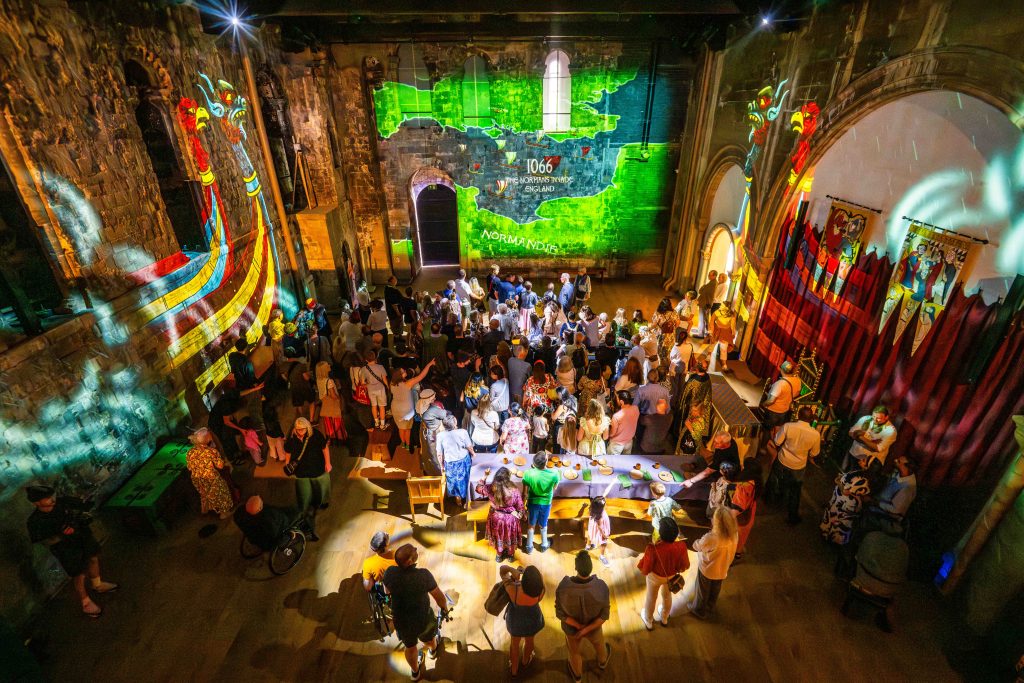 Visitors inside the Great Hall of Norwich Castle Keep watching an immersive projection show. Brightly lit images of Viking ships and a green map of Normandy and England (dated 1066) are projected onto the stone walls. The hall is filled with people of all ages, some in wheelchairs, gathered around long tables and historic displays, with banners hanging on the walls.