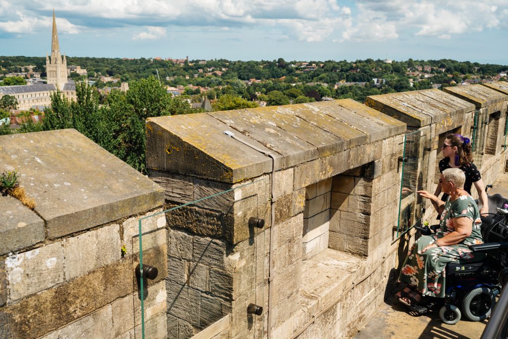 Two wheelchair users on the battlements of Norwich Castle Keep, looking out over the city skyline. One person points while the other observes through a glass viewing panel. In the distance, Norwich Cathedral’s tall spire rises above a mix of trees and buildings.