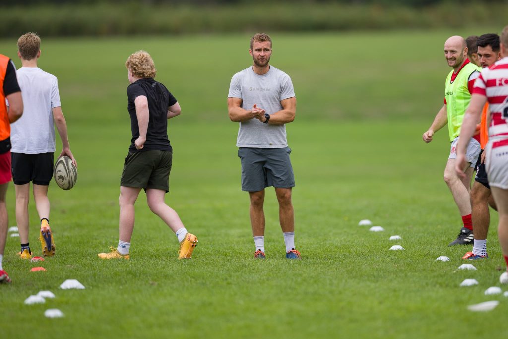 A rugby coach stands on a grassy field clapping his hands, surrounded by players during a training session. One player holds a rugby ball while others in sports kit and bibs smile and prepare for drills, with cones marking the practice area.