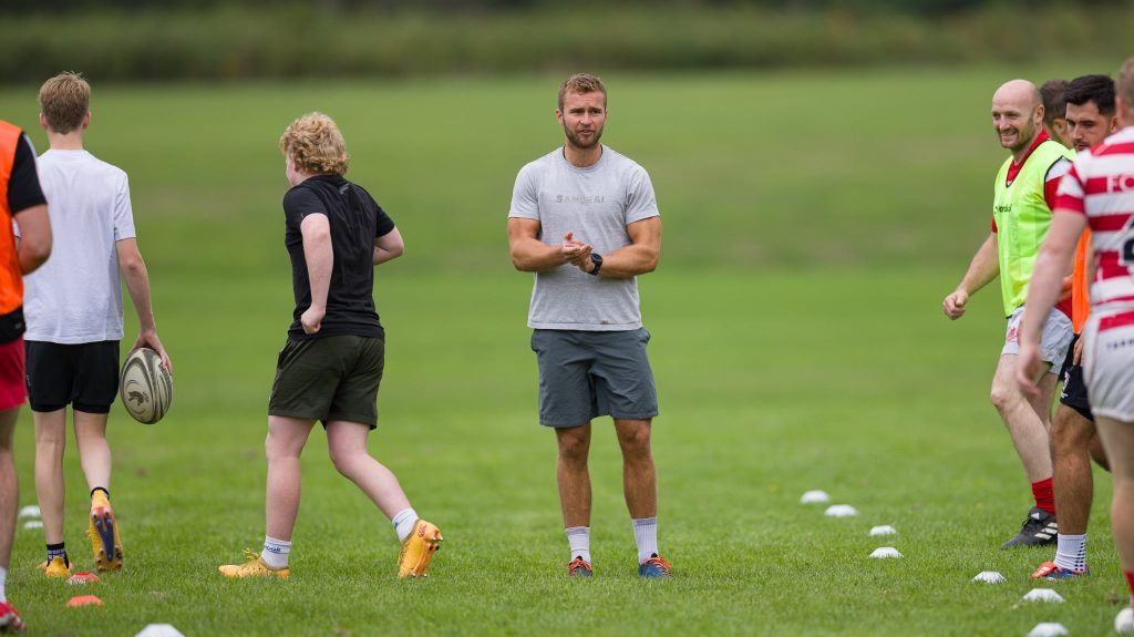 A rugby coach stands on a grassy field clapping his hands, surrounded by players during a training session. One player holds a rugby ball while others in sports kit and bibs smile and prepare for drills, with cones marking the practice area.