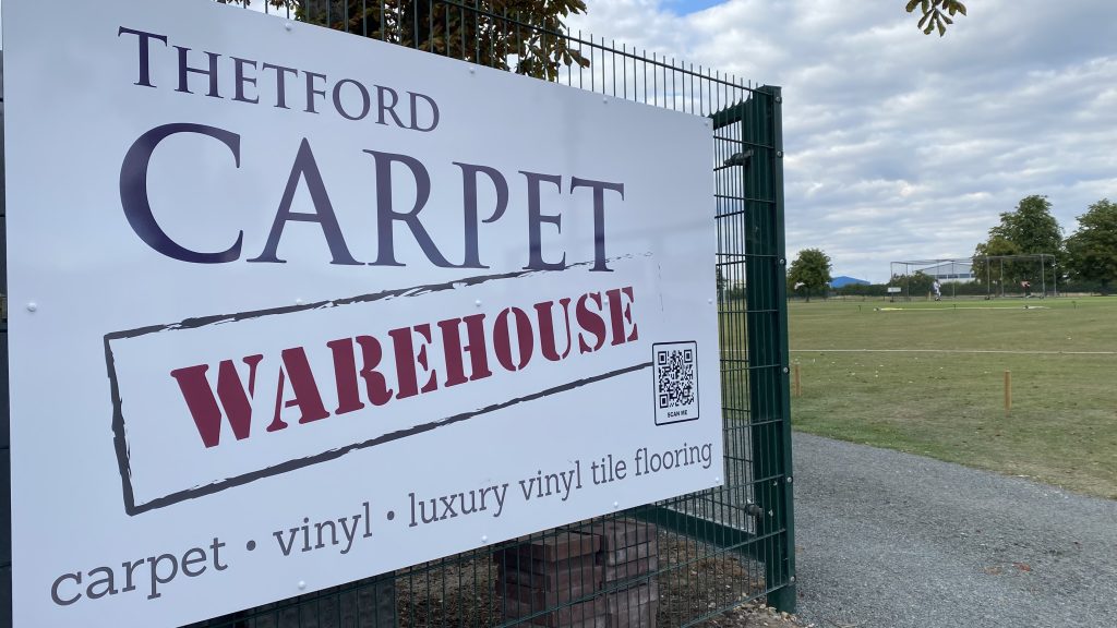 Large outdoor sign attached to a metal fence reads: “Thetford Carpet Warehouse – carpet • vinyl • luxury vinyl tile flooring.” A QR code is included at the bottom right. Behind the fence is an open grassy sports field with goal nets and trees.