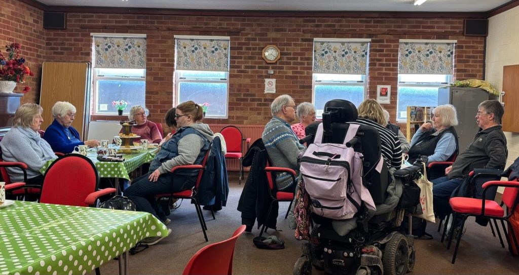 A group of people sit chatting around tables covered in green polka-dot tablecloths at a community centre. Some are enjoying tea and cake, while others are engaged in conversation. The room has brick walls, floral window blinds, and a welcoming, informal atmosphere, suggesting a friendly social gathering or community coffee morning.