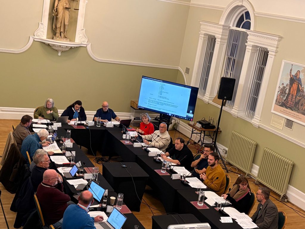 A group of councillors and officers sit around a large U-shaped table during a formal meeting in a high-ceilinged room with pale green walls and tall arched windows. Laptops, papers, and nameplates are spread across the tables, and a large screen displays an agenda on the wall. A statue and historical artwork decorate the room. The atmosphere is serious and focused as members discuss local matters, including the car park proposal.