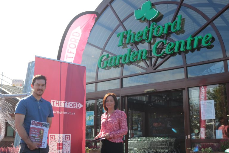 Two people stand outside Thetford Garden Centre next to a large red “For Thetford” banner, holding information leaflets.