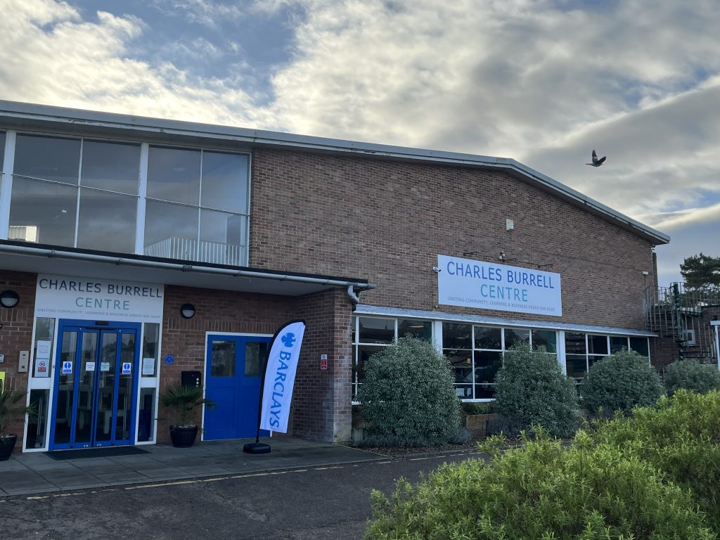 Exterior of the Charles Burrell Centre in Thetford, a red-brick community building with blue doors and signage. A Barclays-branded flag stands outside the entrance under a cloudy sky.