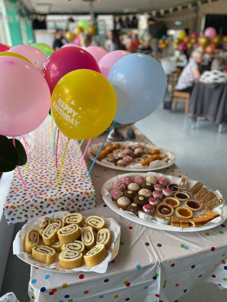 Close-up of a decorated party table with colourful “Happy Birthday” balloons and platters of cakes, jam tarts, and mini rolls. In the background, people are gathered and chatting in a festively decorated room.