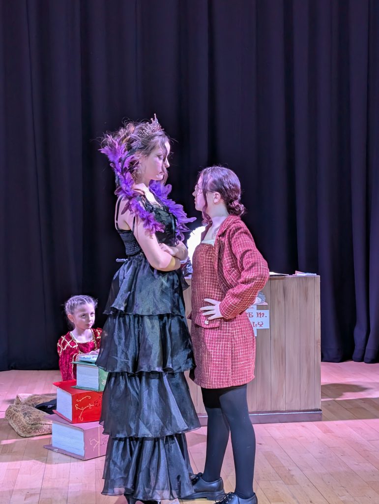 Three children perform a dramatic theatre scene. One wears a long black tiered dress, tiara, and purple feather boa, facing another dressed in a red checked jacket. A third child sits nearby in a red costume beside large decorative books.