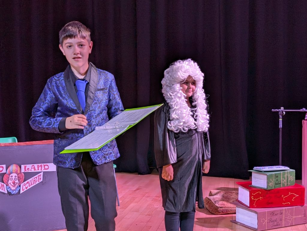 Two children perform on stage during a play. One wears a blue patterned jacket and tie while holding a script, and the other wears a curly white judge’s wig and dark robe. Behind them, stage props include a desk labeled “Wonderland Court” and a stack of oversized books.
