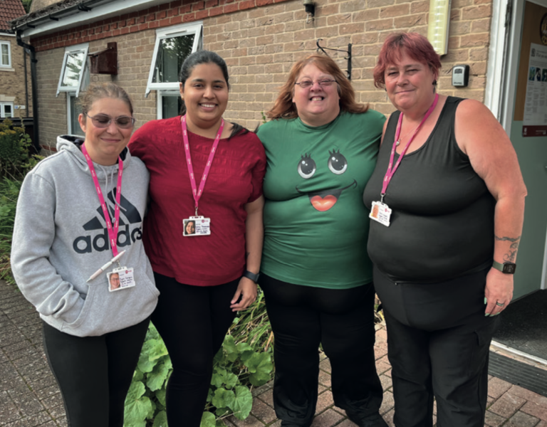Four smiling women stand together outside a brick building, wearing name badges on pink lanyards. One is dressed in a grey Adidas hoodie, another in a red top, one in a green T-shirt with a cartoon face design, and another in a black sleeveless top. They appear to be part of a care or support team, standing close together and posing happily for the photo.